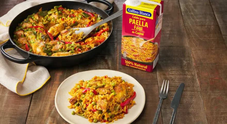 Paella de carne recién hecha en sartén y plato, lista para disfrutar. Un caldo Gallina Blanca para paella fácil de carne a su lado.