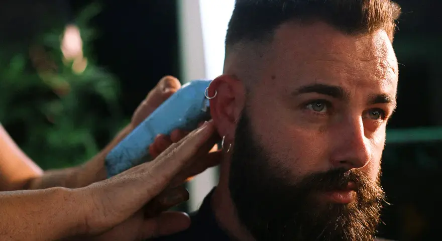 Un hombre con barba y aros en las orejas recibe un corte de pelo moderno, anticipando las tendencias de corte de pelo hombre 2026.