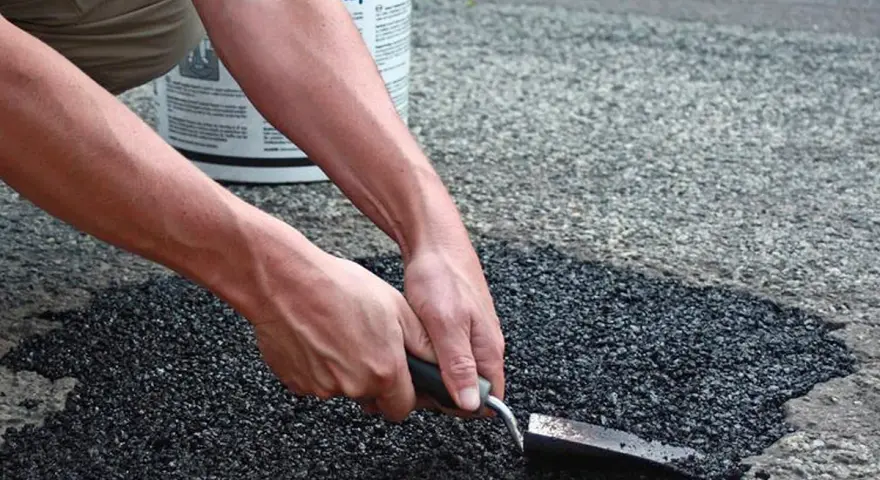 Händer som reparerar asfalt med en spade. En hink med "Asphalt" syns i bakgrunden.