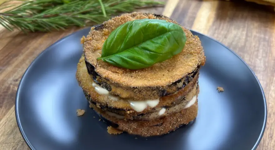 Deliziose cotolette di melanzane impilate, farcite con formaggio cremoso e guarnite con una foglia di basilico fresco.