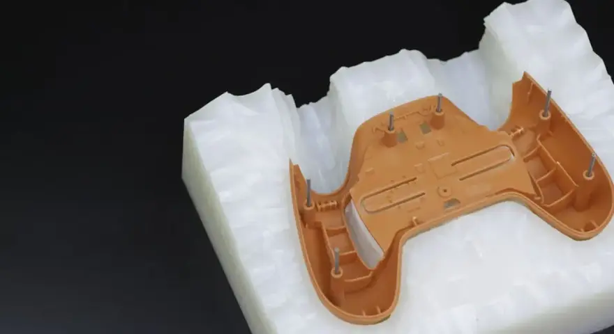 A brown plastic part, likely from a game controller, is placed on a white, textured material. This setup suggests a step in the polyurethane vacuum casting process, where a mold is being prepared.