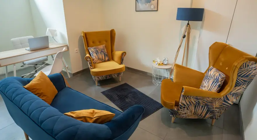 Nowoczesny gabinet psychologiczny z żółtymi fotelami, niebieską kanapą i biurkiem.