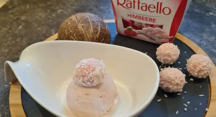 Ein Löffel Raffaello Himbeer Eis in einer Schale, daneben drei Raffaello-Kugeln und eine Kokosnuss.