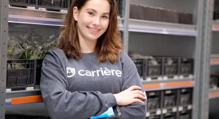 Young woman in a grey sweatshirt with "carrière" logo, holding a scanner, in a warehouse with shelves of black crates.