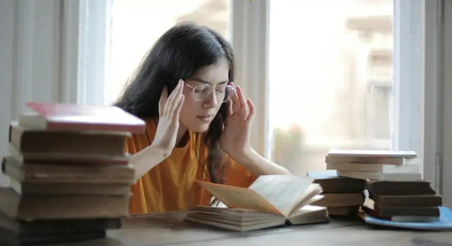 Ragazza con occhiali e mani sulle tempie, circondata da libri. Potrebbe soffrire di mancanza di concentrazione cause.