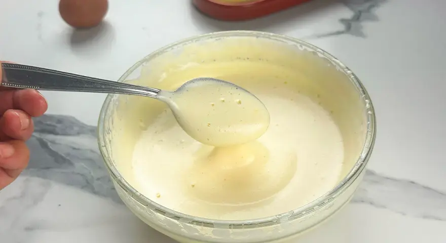 Préparation d'un tiramisu : une cuillère de pâte à bombe aérienne est prélevée d'un bol en verre. Un œuf et des biscuits à la cuillère sont visibles.