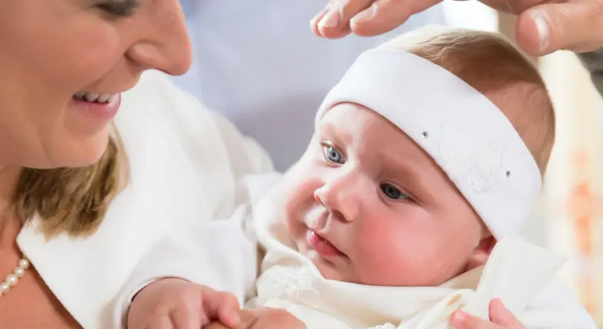 Uśmiechnięta mama trzyma dziecko w białej szatce. Dłoń ojca błogosławi malucha. Zastanawiasz się, ile kosztuje chrzest?
