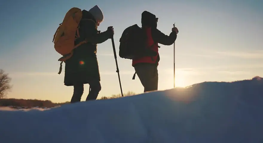 Dwie osoby z plecakami i kijkami trekkingowymi idą przez ośnieżony teren. W tle zachodzące słońce, przypominające czerwone wierchy szczyty.