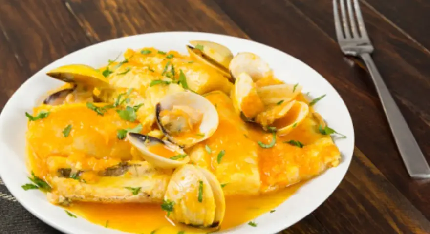 Merluza a la marinera con almejas, servida en un plato blanco sobre una mesa de madera oscura.