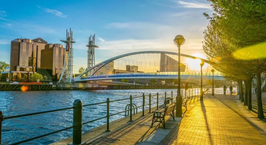Promenade ensoleillée au bord de l'eau, avec un pont moderne et des bâtiments reflétant le soleil. Idéal pour visiter Manchester ou Liverpool.