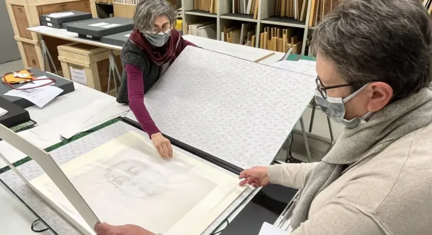 Deux personnes manipulent un dessin au crayon, peut-être un portrait, sur une table. Le vernis protège l'œuvre.