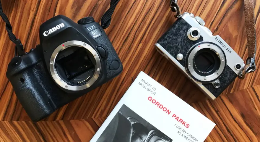 Dwa aparaty, Canon EOS 6D Mark II i Olympus PEN, leżą obok książki Gordona Parks'a "Aparat to moja broń". Pełna klatka i mniejszy aparat gotowe do akcji.