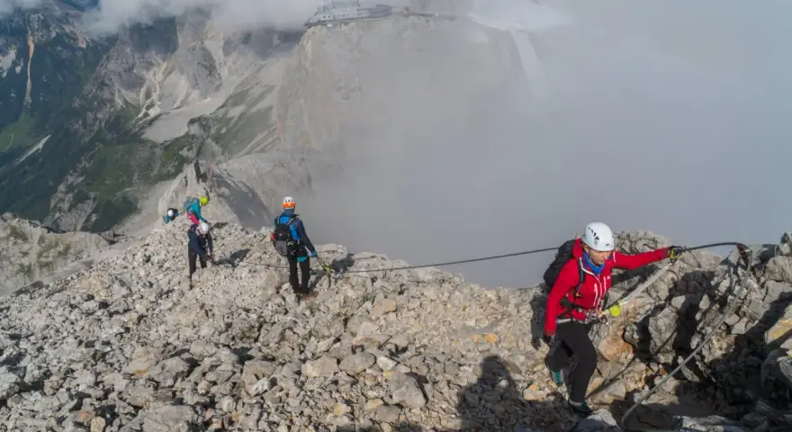 Grupa turystów pokonuje via ferrata polska. Wspinają się po skalistym grzbiecie, asekurowani linami. W tle widać góry i budynek na szczycie.