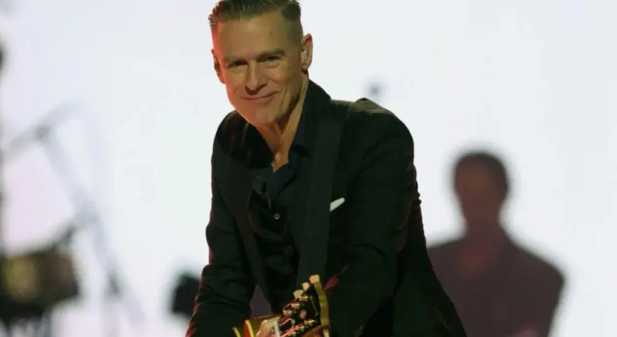 Bryan Adams w czarnym garniturze gra na gitarze. Sprawdź jego nadchodzące wydarzenia i koncerty.