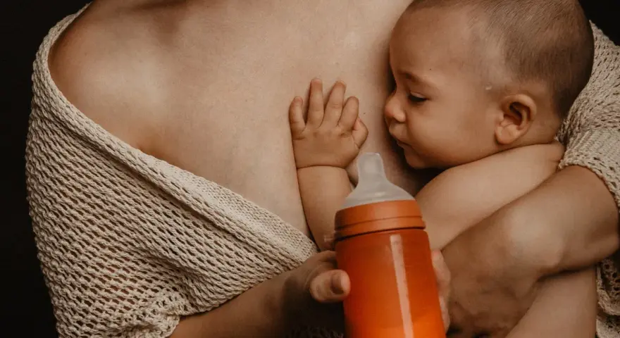 Bebé succionando un biberón naranja, con la mano apoyada en el pecho de la madre. Se aprecian varios tipos de tetinas en el mercado.