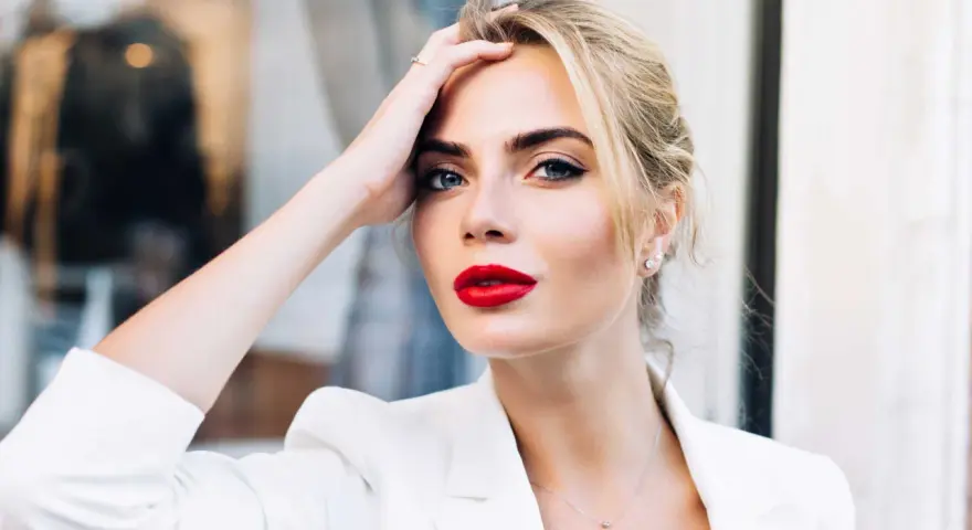 Frau mit roten Lippen und weißem Blazer, Hand am Kopf.