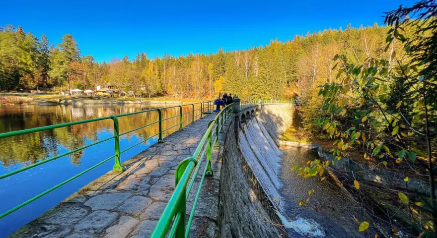 Kamienna zapora z zieloną balustradą, wodospadem i jesiennym lasem w tle.