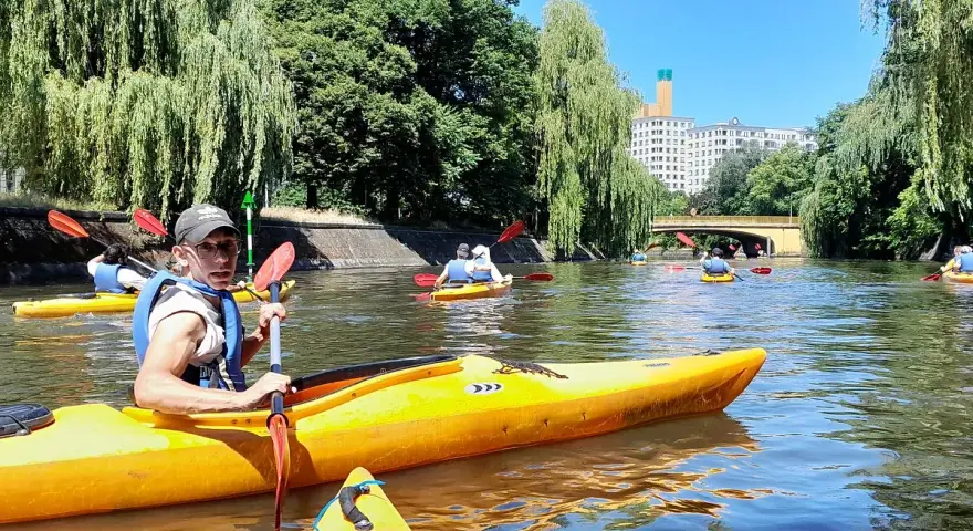 Eine Gruppe von Menschen paddelt auf gelben Kajaks auf einem Fluss in Berlin. Bäume säumen das Ufer, im Hintergrund ist ein Gebäude zu sehen.