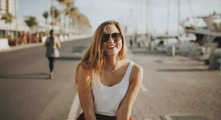 Glückliche junge Frau mit Sonnenbrille und weißem Top genießt ihr Praktikum in Spanien am Hafen.