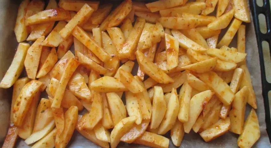 Gotowe do pieczenia frytki w piekarniku, pokrojone w łódeczki i doprawione przyprawami, leżą na blasze wyłożonej papierem.