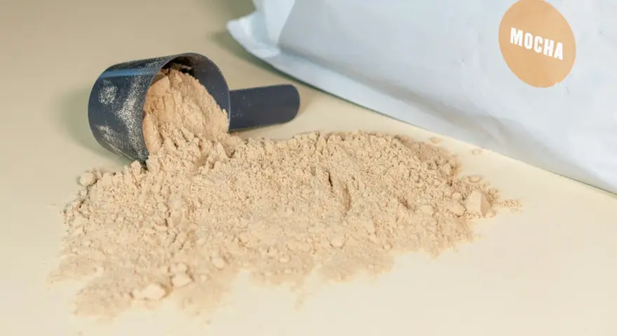 Moccha-smakande proteinpulver spillt från en skopa, med påsen i bakgrunden. Perfekt för att hålla koll på proteinpulver kcal.