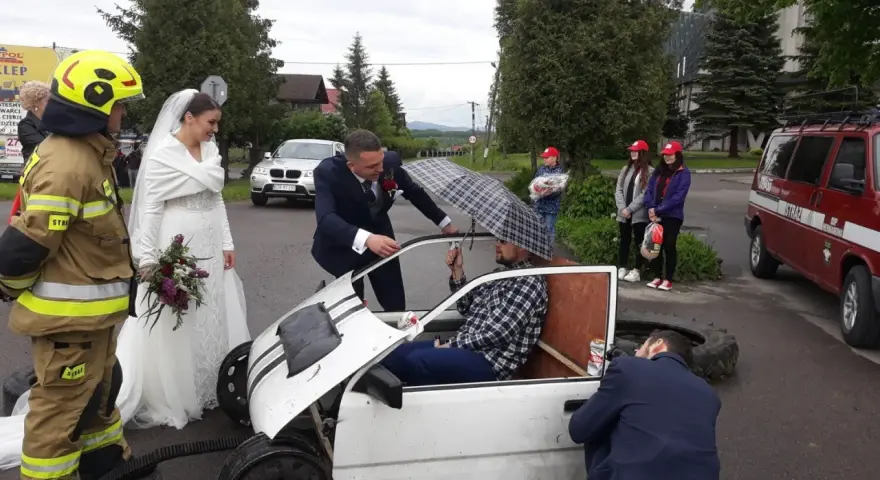 Brama weselna z udziałem strażaka, pana młodego i panny młodej. Pan młody rozmawia z druhem siedzącym w prowizorycznym samochodzie.