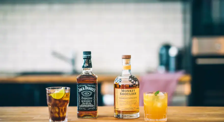 Zwei Flaschen Whiskey, Jack Daniel's und Monkey Shoulder, stehen neben zwei Cocktails auf einem Holztisch.