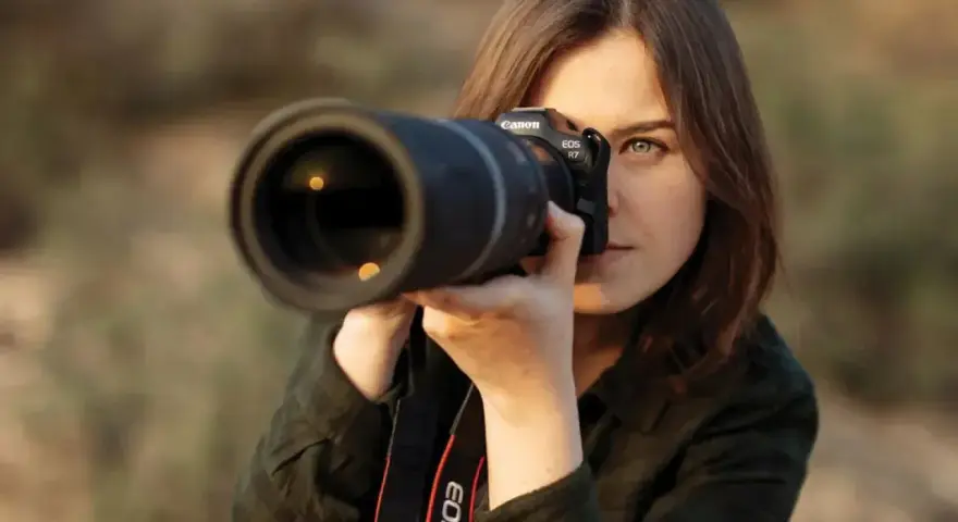 Kobieta z aparatem Canon EOS R7, skupiona na kadrze. Jej oczy błyszczą, a obiektyw sugeruje zaawansowane techniki fotograficzne.