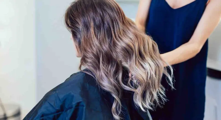 Kobieta z falowanymi włosami w odcieniach brązu i subtelnym balejażem blond. Fryzjerka poprawia jej fryzurę.