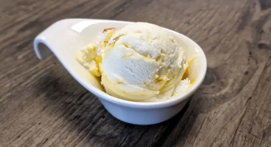 Ein Löffel cremiges Buttermilch-Eis, beträufelt mit Honig, in einer weißen Schale auf Holztisch.