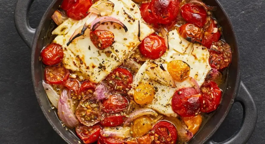 Heißer, gebackener Feta mit Kirschtomaten und roten Zwiebeln in einer Pfanne, mit Kräutern bestreut.