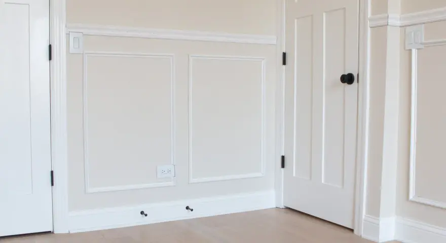 A room with a DIY chair rail, wainscoting, and white doors. The walls are painted a light cream color.