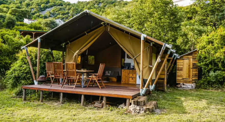 Glamping w Czarnogórze: namiot z drewnianym tarasem, stołem i krzesłami, otoczony zielenią. Idealne miejsce na relaks.