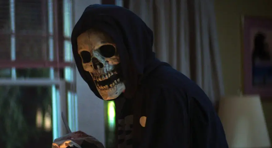 Eine Person mit Totenkopfmaske und Kapuze hält blutige Gegenstände.