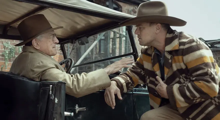 Due uomini in auto discutono animatamente. Uno, al volante, con cappello e giacca chiara, sembra implorare l'altro. Quest'ultimo, con un cappello simile e una giacca a righe, ascolta con espressione tesa, forse coinvolto negli eventi del "killer of the...
