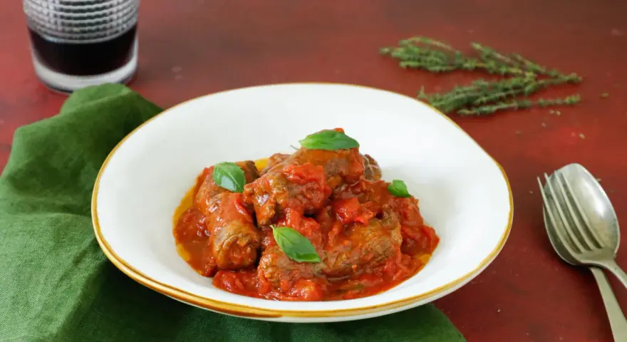 Involtini di carne in padella con sughetto, guarniti con basilico fresco, serviti in un piatto bianco su un tovagliolo verde.