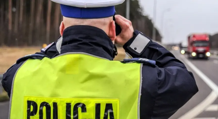 Policjant w kamizelce odblaskowej obserwuje drogę, gdzie widać ciężarówkę. Na drodze obowiązuje zakaz wyprzedzania dla ciężarówek.