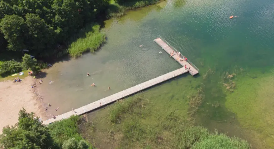 Letnie popołudnie nad jeziorem Hańcza. Ludzie kąpią się i odpoczywają na plaży, inni spacerują po drewnianym pomoście.