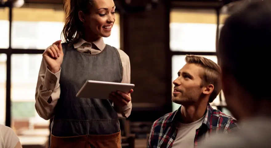 Camarera sonriente atiende a clientes de restaurante, tomando nota en una tablet.