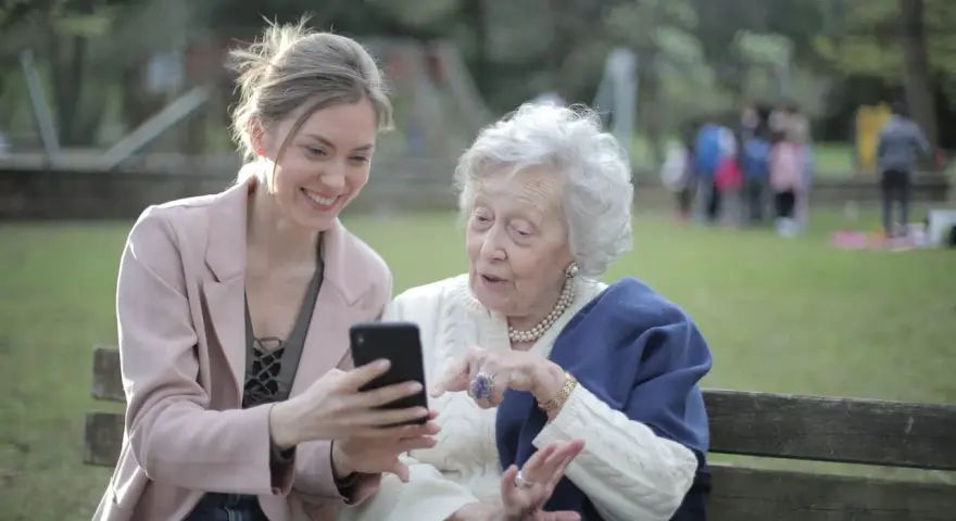 Młoda kobieta pokazuje coś na telefonie starszej pani. To scena z życia, jak z kursu opiekuna osoby starszej, gdzie uczymy się wspierać seniorów w codzienności.