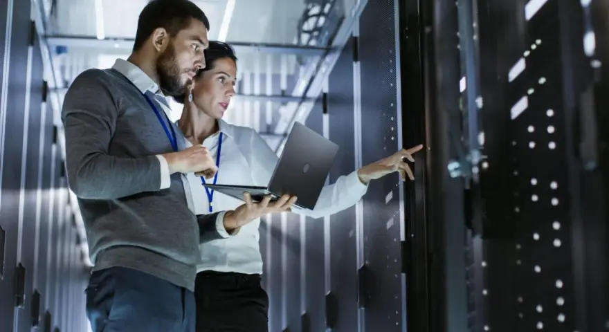 Two IT professionals collaborate on network management, one holding a laptop while the other points to a server rack.