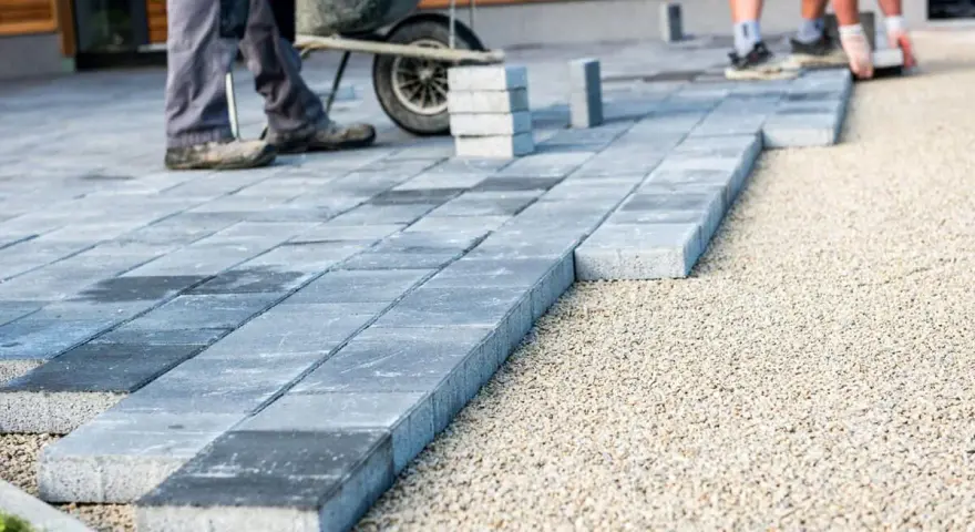 Układanie płyt betonowych na podjeździe – alternatywa dla kostki. Pracownicy budowlani układają szare płyty na podsypce żwirowej.