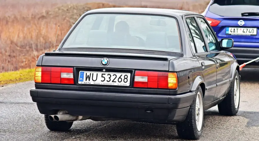 Tył czarnego BMW E30 z tablicą rejestracyjną WU 53268, holowanego przez niebieskiego SUV-a.