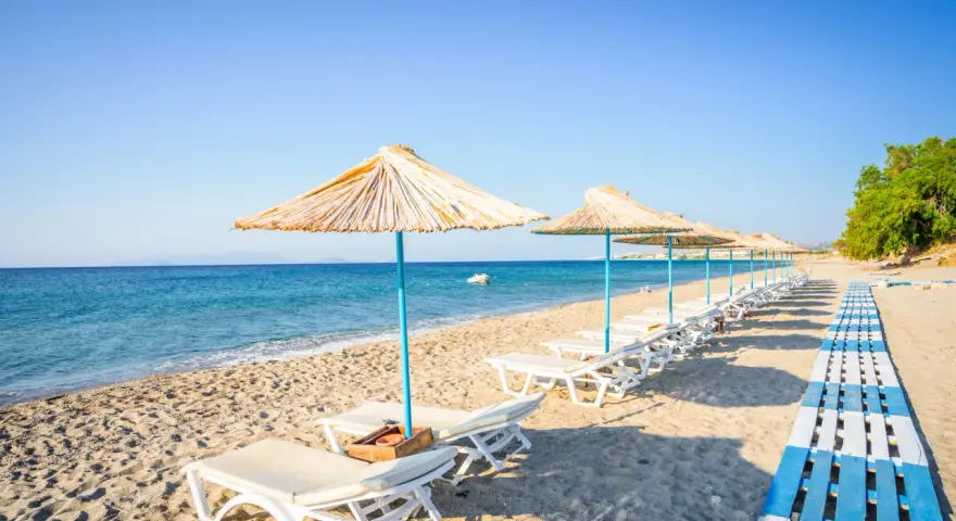 Plaża na wyspie Kos. Rzędy leżaków pod słomianymi parasolami, drewniana ścieżka i spokojne morze.