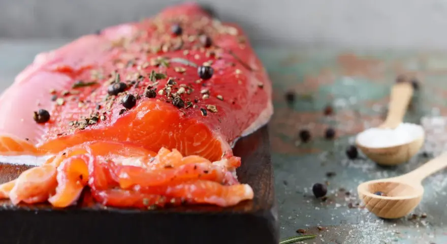 Un filet de gravlax de truite, assaisonné de poivre et d'herbes, prêt à être dégusté. Deux cuillères en bois contiennent sel et genièvre.