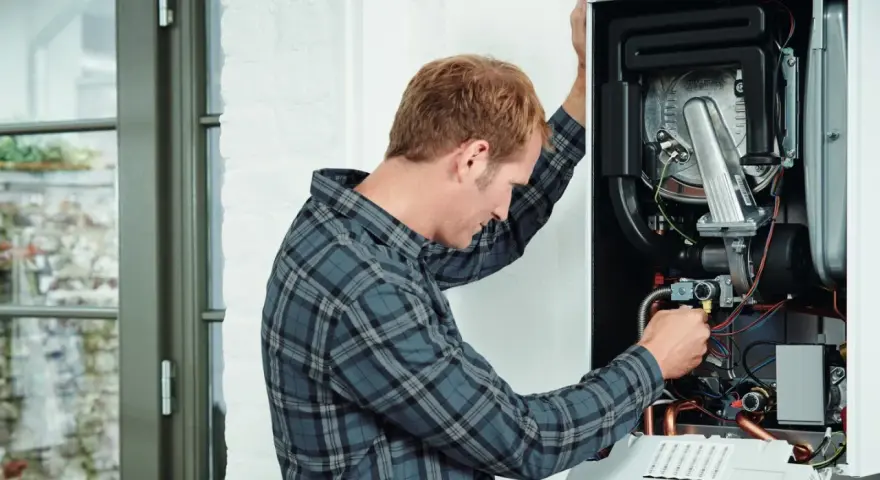 Tecnico che controlla lo scambiatore di calore della caldaia, con cavi e tubi visibili all'interno.