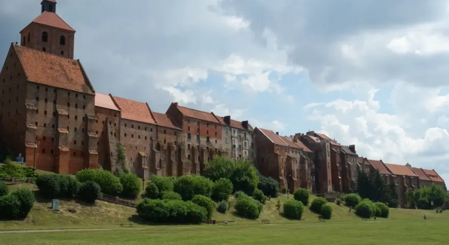 Długi, ceglany zamek w Grudziądzu w jeden dzień. Zielona trawa i krzewy przed zabytkową budowlą.