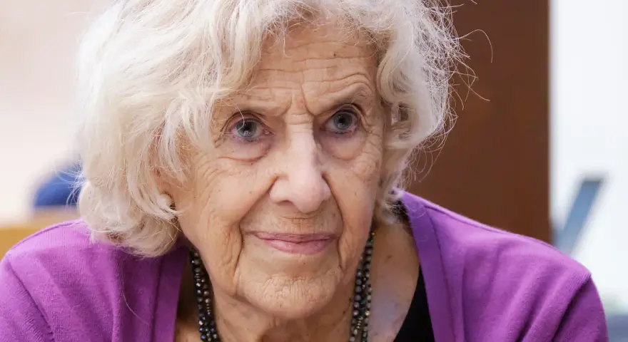 Retrato de una anciana con cabello blanco y ojos azules, luciendo un suéter morado. Su rostro arrugado evoca la sabiduría y el significado de una vida plena, como el nombre Manuela.