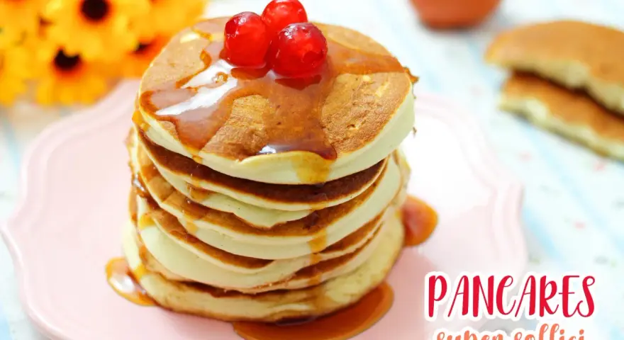 Pancakes con albumi, soffici e dorati, guarniti con sciroppo e ciliegie rosse, su un piatto rosa.