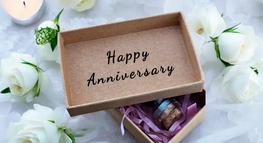 Otwarta szkatułka z napisem "Happy Anniversary" i małą buteleczką w środku, otoczona białymi różami i płonącą świecą. To idealny prezent na 4 rocznicę ślubu.